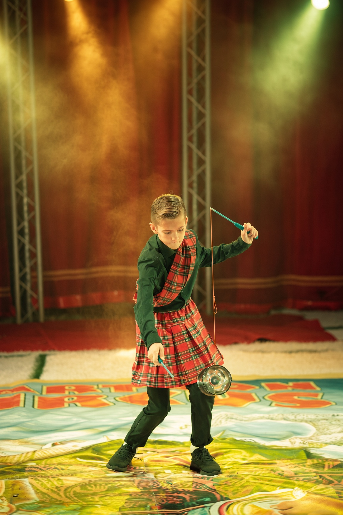 Junge in rot-grünem Kilt spielt mit einem Diabolo in der Zirkusmanege vor rotem Bühnenvorhang