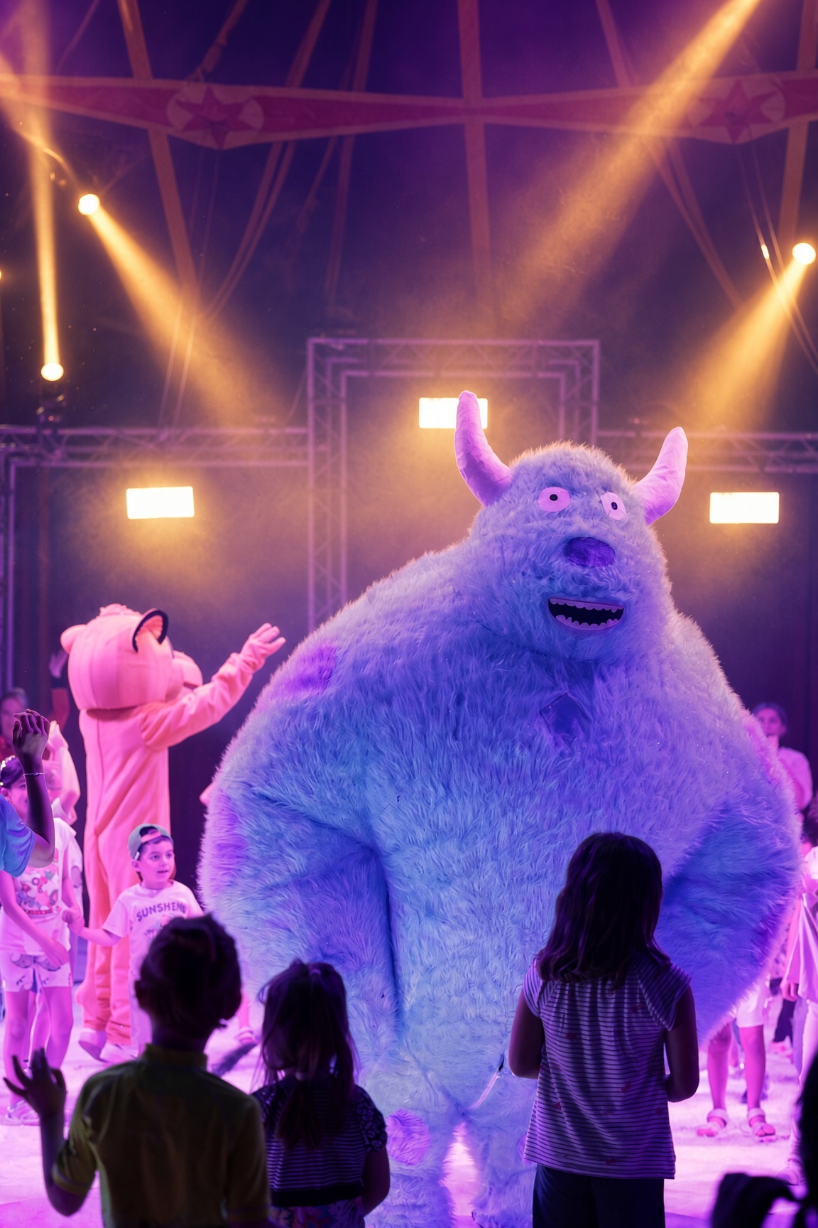 Ein blaues Plüschmonster tanzt mit Kindern unter hellem Scheinwerferlicht im Zirkuszelt